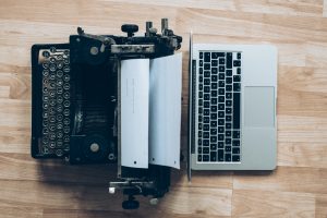Auf einem Holzboden stehen sich links eine Schreibmaschine mit eingestecktem Papier und recht ein Macbook gegenüber